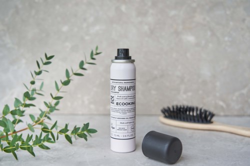 A bottle of dry shampoo, black hairbrush, and green leaves on a gray surface.