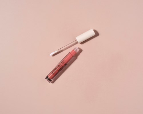 A lip gloss tube with an open applicator lies on a pink surface.