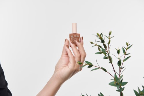 A hand holding a beige nail polish bottle next to a green leafy plant.