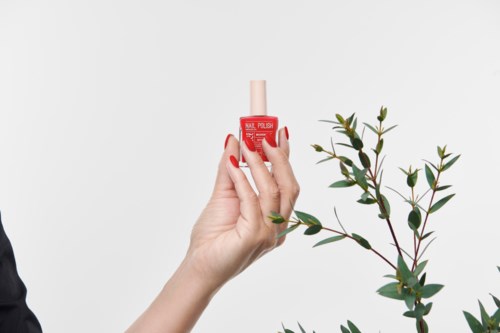 A hand holds a red nail polish bottle near some green leaves on a plain white background.