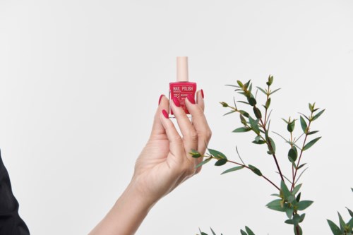A hand holds a pink nail polish bottle near green plant leaves against a white background.