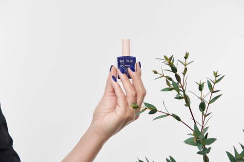 A hand holds a blue nail polish bottle near green leafy branches on a plain background.