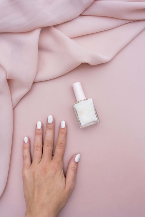 A hand with white nails resting on pink fabric next to a bottle of white nail polish.