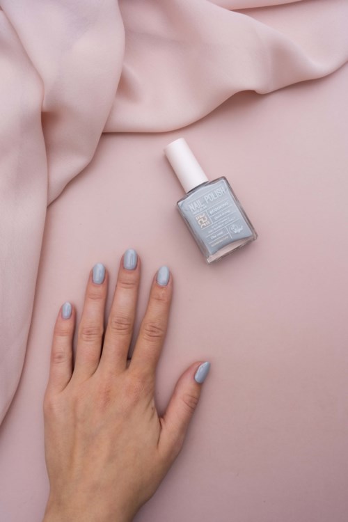 A hand with light blue painted nails is shown next to a bottle of silver nail polish on pink fabric.