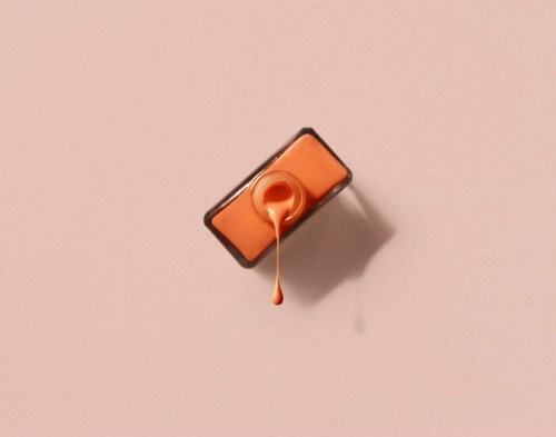A nail polish bottle with orange polish and a brush, against a pink background.