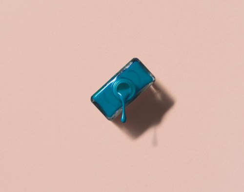 A small bottle of blue nail polish spilling onto a pink surface. The shadow is visible.