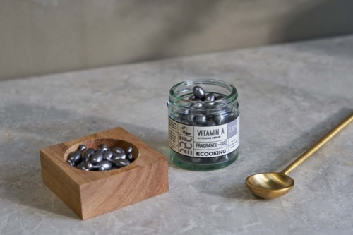 Lille glas med sølvfarvede vitamin A-kapsler, trækasse med kapsler, guldske på grå overflade.