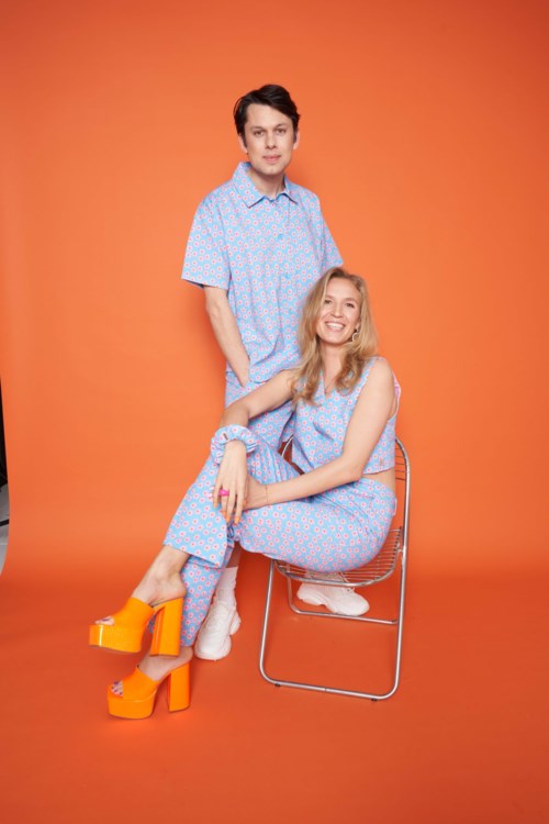 A young man and woman in matching blue clothes with colorful patterns, set against an orange background.