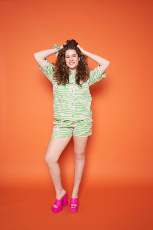 A young woman with curly hair in fun green and white striped clothing, smiling, standing in front of an orange wall.