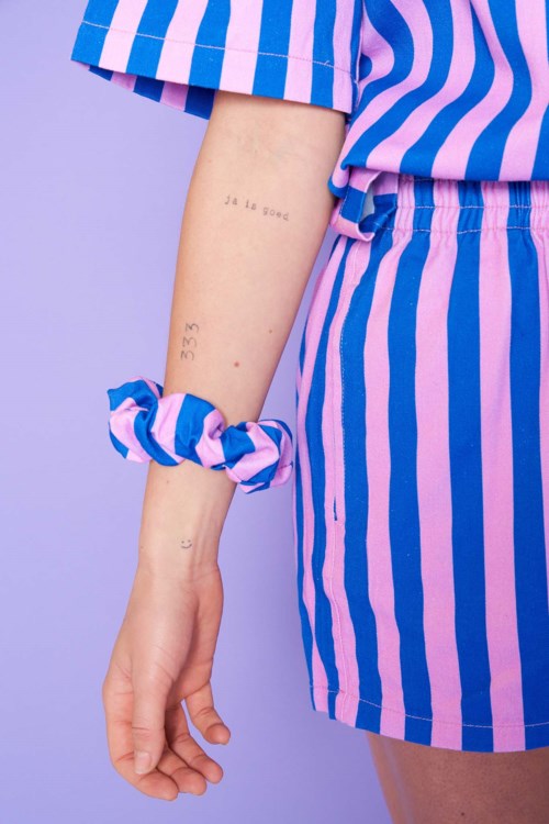 Person wears a blue and pink striped outfit, has tattoos on arm, and a scrunchie on wrist against purple background.