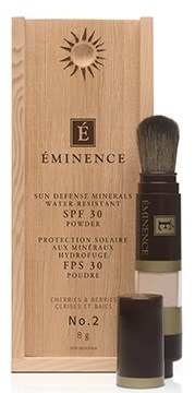 En trälåda innehåller en flaska Eminence solskydd, med en borste och lock bredvid. Boxen har en solsymbol och text.