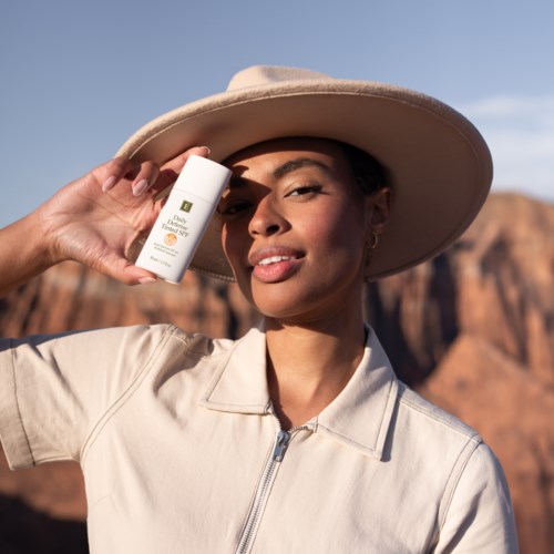 A woman outdoors, holding a sunscreen bottle, wearing a wide-brimmed hat, against a desert landscape.