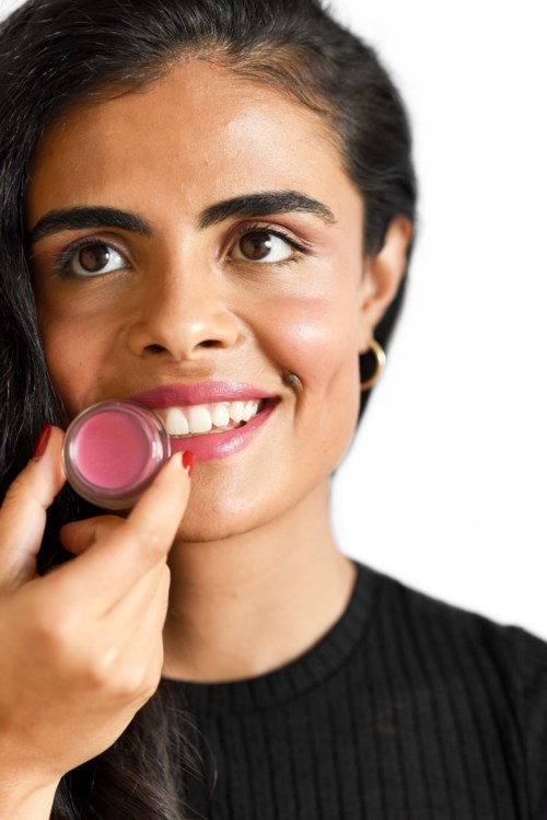 A woman with dark hair applying pink lipstick, smiling, wearing a black top, on a white background.