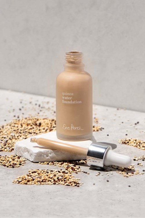 A beige bottle labeled 'quinoa water foundation' and a dropper lie on a white surface with seeds around.