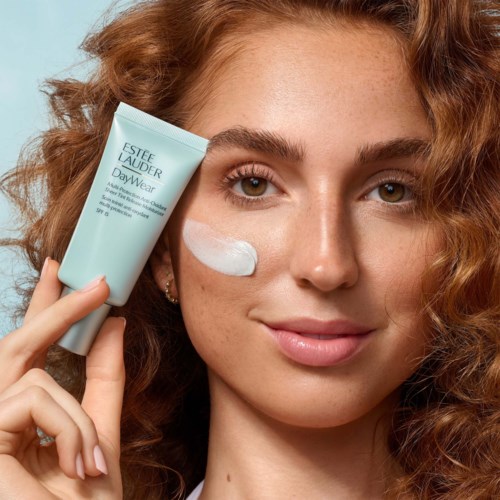 Eine Frau mit lockigem roten Haar trägt Creme auf die Wange auf und hält eine blaue Tube mit Estée Lauder Hautpflege.