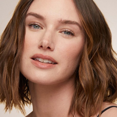 A woman with shoulder-length brown hair and fair skin, wearing light makeup and a dark strap top.
