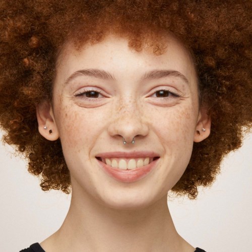 A young woman with curly red hair, freckles, and a nose piercing, smiling at the camera.