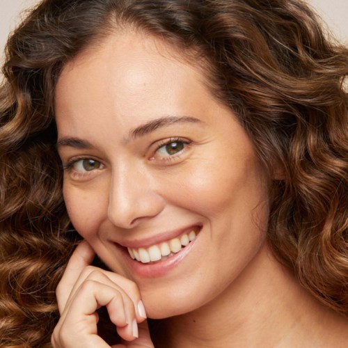 Eine Frau mit lockigem Haar lächelt sanft, berührt ihr Gesicht vorsichtig, mit freundlichem Ausdruck und leuchtenden Augen.