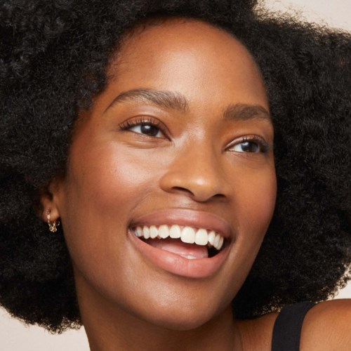 A smiling woman with curly hair and earrings, showing her teeth and sticking her tongue out playfully.