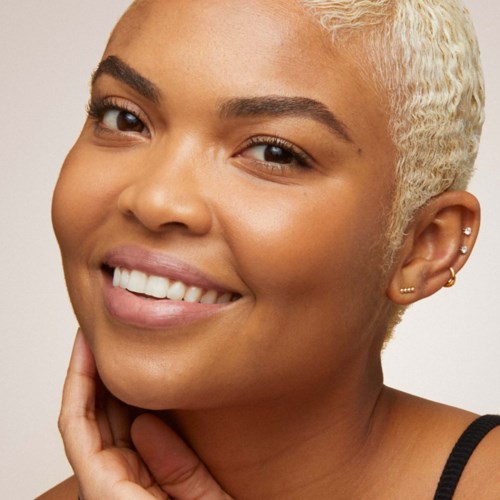 Eine Frau mit kurzen, platinblonden Haaren, die sanft lächelt, ihre weißen Zähne zeigt und kleine Ohrringe trägt.