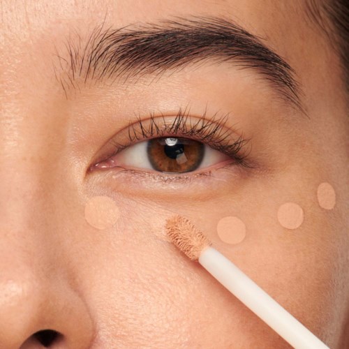En person applicerar beige concealer under ögat med en tops eller borste. Huden ser jämn och ljus ut.
