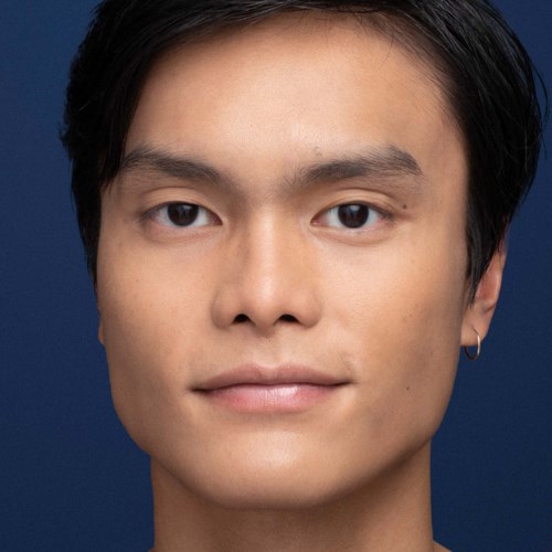A young man with short black hair and a gold earring, smiling slightly against a blue background.