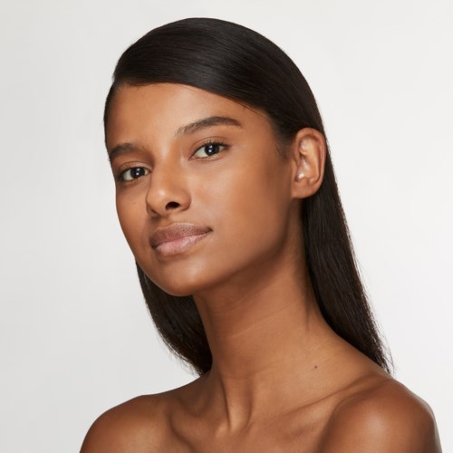Eine junge Frau mit glatten, dunklen Haaren und klarer Haut schaut selbstbewusst in die Kamera. Sie trägt keinen Schmuck und hat einen neutralen Hintergrund.