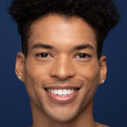 Ein lächelnder junger Mann mit kurzen, lockigen Haaren, Ohrringen und dunkelblauem Hintergrund.
