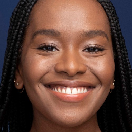 Smiling young woman with braided hair and earrings, close-up shot against a dark background.