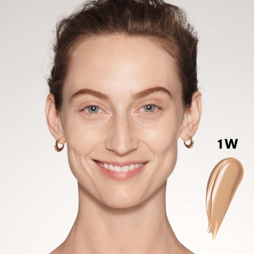 En leende kvinna med klar hy och blå ögon, visar en makeupprovning märkt '1W' på kinden.