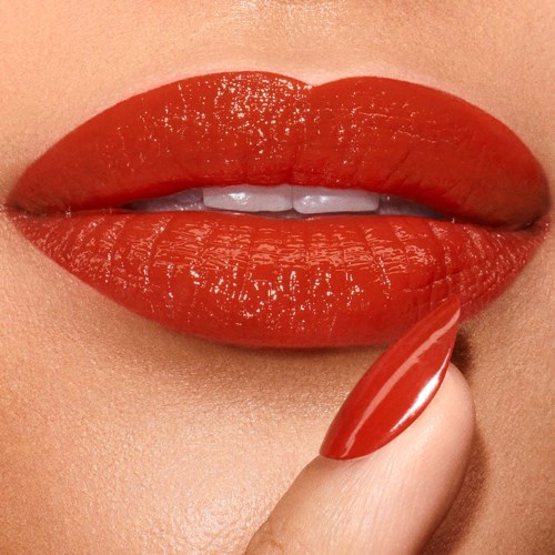 Close-up of lips with glossy red lipstick, with a finger touching the lip. The lips are slightly parted, revealing teeth.