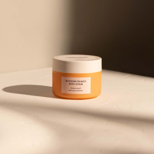 En krukke med Blossom Crunch Body Scrub i oransje farge, hvitt lokk og etikett. Den står på en hvit overflate med skygger.