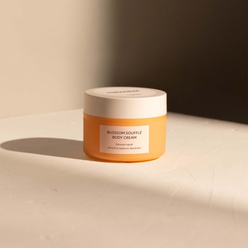 En liten burk Blossom Soufflé Body Cream med vit lock och orange behållare. Den står på en vit yta.