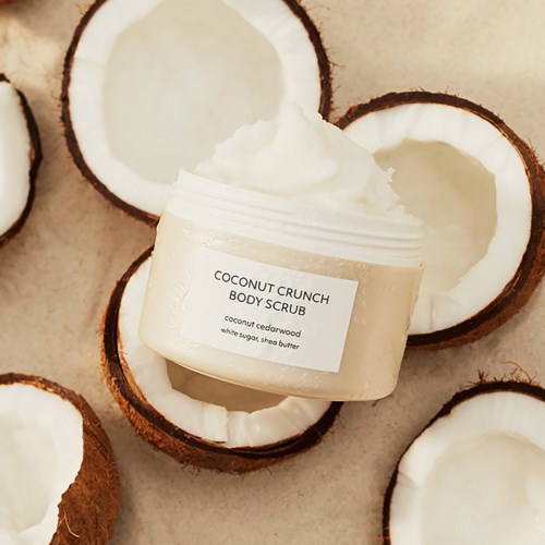 A jar of coconut body scrub surrounded by halved coconuts with white flesh. The label reads 'Coconut Crunch Body Scrub'.