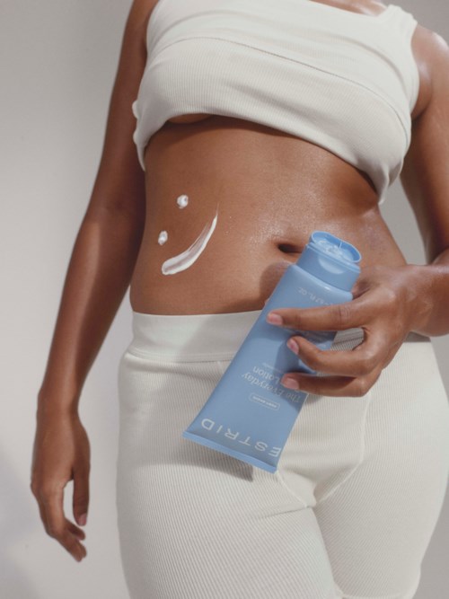 En kvinna smörjer in magen, håller en blå flaska, bär en vit crop top och byxor.