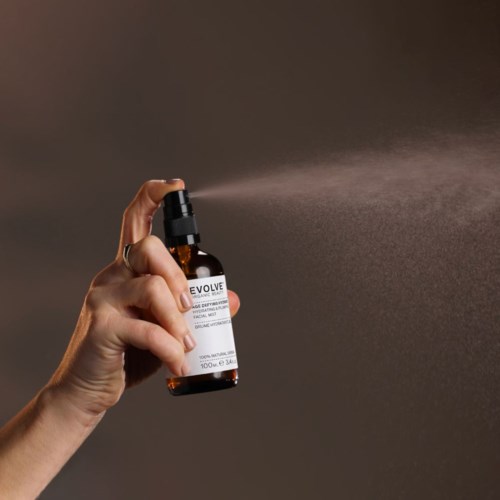 A hand sprays a liquid from a small brown bottle with a pump onto skin. The background is plain and dark.