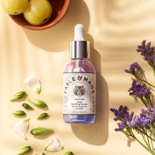 A pink and purple glass bottle of Fable & Mane Amla soothing serum with a white dropper cap, surrounded by flowers.