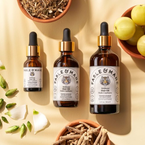 Three brown glass bottles with black and gold dropper caps labeled Fable & Mane hair oil, surrounded by green apples, white flowers, and herbs.