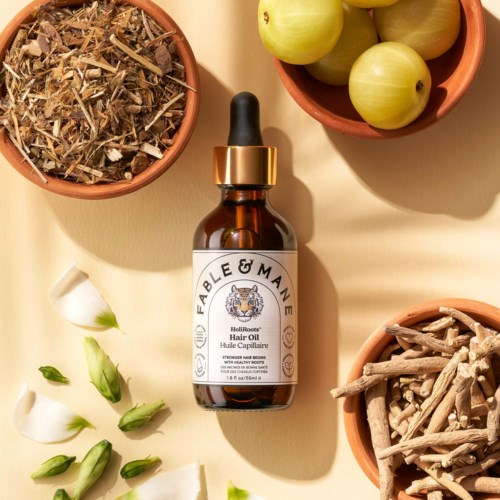 A brown glass bottle of Fable & Mane hair oil with a dropper cap, surrounded by bowls of herbs, green apples, and roots.