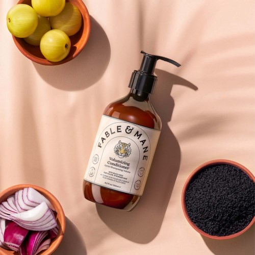 A bottle of Fable & Mane conditioning hair product surrounded by bowls of onions, black powder, and green fruits.