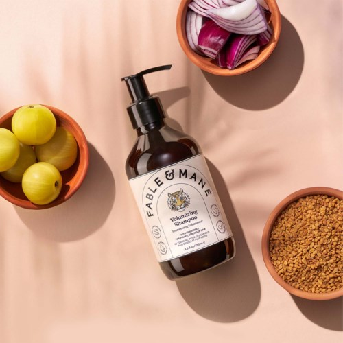 A brown shampoo bottle with a pump, labeled FABLE & MARE, surrounded by bowls of apples, purple petal fragments, and grains.