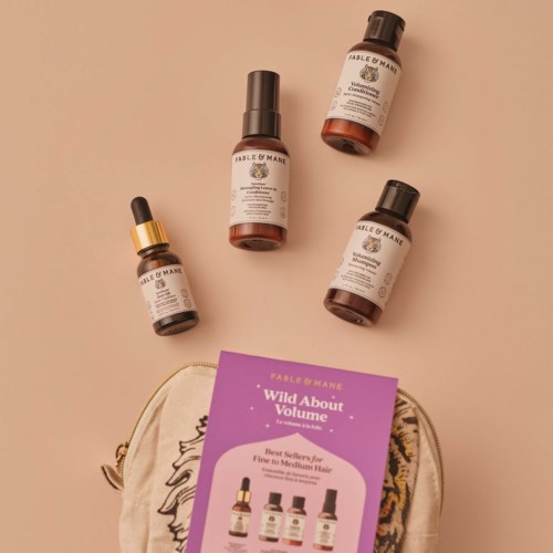 Three brown bottles of Fable & Mane hair products with black caps, a purple booklet titled Wild About Volume, and a beige bag with a colorful leopard print.