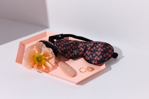 En rosa tallerken holder en blomsteraktig sovemaske, en blomst, en ring og en lotionflaske. Oppsettet er enkelt og ryddig.