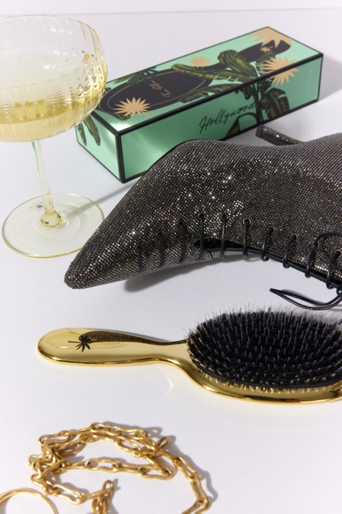 Een glas witte wijn, een versierde doos, een zwart textuurkussen, een gouden haarborstel en gouden sieraden op een witte ondergrond.