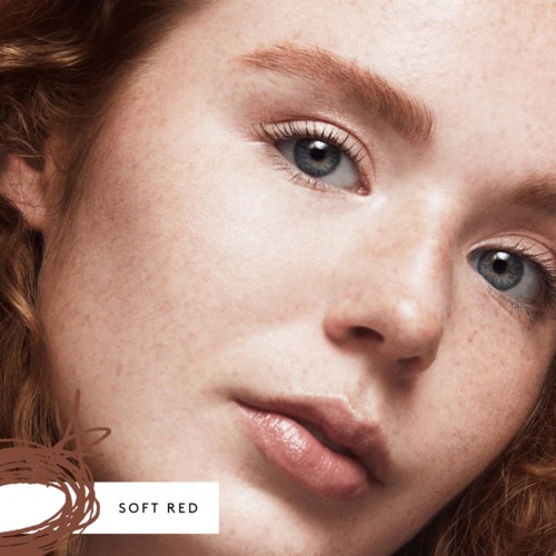 Närbild av en ung kvinnas ansikte, ljus hy med fräknar, blå ögon och naturlig makeup, mjukt rött hår.