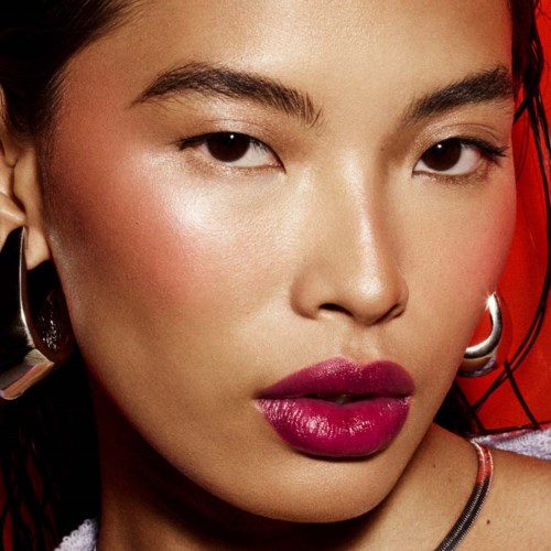 Close-up of a woman with bold makeup, dark hair, earrings, and a vivid red background.