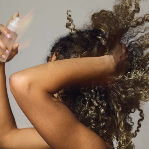 A person with curly hair sprays hair product, lifting their arm to style or treat their hair.