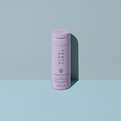 En rosa flaska med Fenty Hair-produkter står upprätt på en ljusblå yta med skugga bakom.