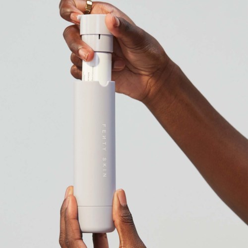 En hand håller en vit cylinderformad flaska märkt 'Fenty Skin' mot en enkel bakgrund.