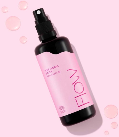 Vaaleanpunaista suihkepulloa, jossa lukee 'Rose Floral Water', ympärillä pieniä vaaleanpunaisia tippoja vaaleanpunaisella taustalla.